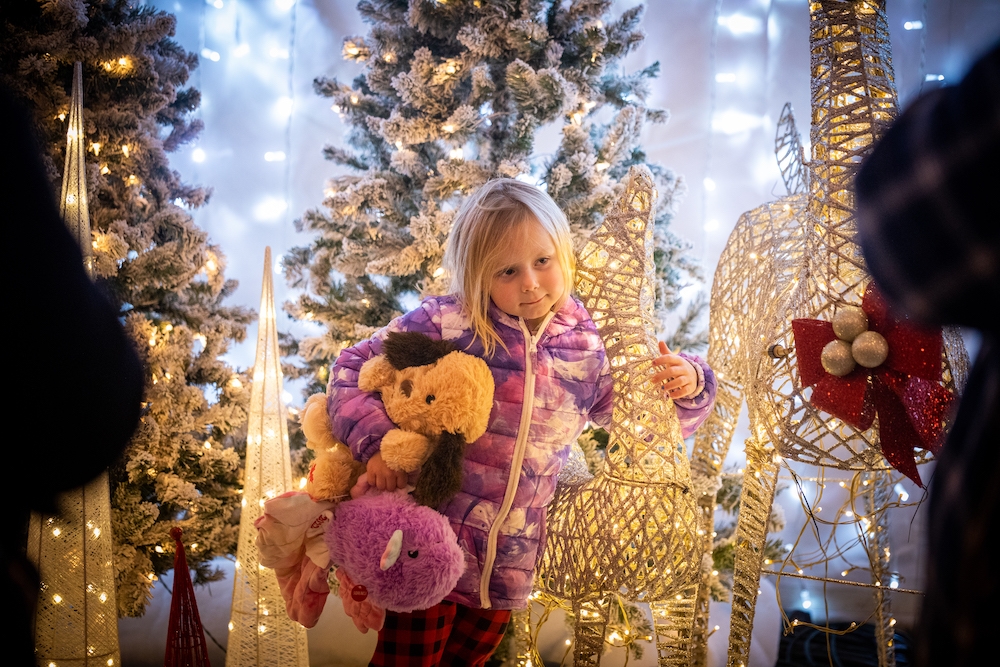 Small girl holding teddy bear with arm around deer decorated in lights